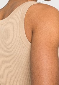 Detailní záběr na rameno a horní část paže osoby v béžovém žebrovaném tílku, který ukazuje texturu pleti a svalový tonus.