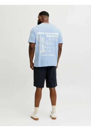 Mann von hinten, der ein hellblaues T-Shirt mit der Aufschrift "Côte Sauvage Bistrot" und einer Café-Szene an der Französischen Riviera trägt, schwarze Shorts, weiße Socken und Schuhe.