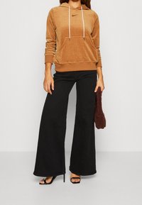 Brun kordflöd jacka med huva och vita dragsko, tillsammans med svarta utsvängda jeans och svarta högklackade sandaler. Vinröd kuvertväska i handen.