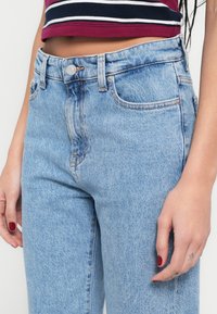 Hele sinised teksapüksid kõrge vöökohaga, viie taskuga ja hõbedase nupuga kinnitusega. Kangal on kerge tekstuur ja tuhmunud viimistlus.