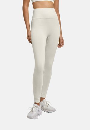 Femme portant des leggings taille haute, sans coutures, blanc cassé et des chaussures de sport blanches, debout devant un fond uni.
