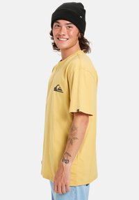 Žluté bavlněné tričko s krátkým rukávem, s černým logem na levém hrudníku. Nositeli trička je na hlavě černá čepice a má viditelné tetování na ruce.