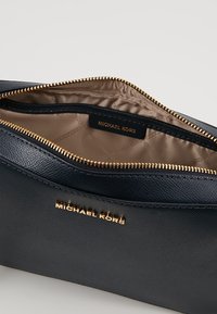 Námornícky kožený pouzdro se zlatým zipem, s hladkým povrchem. Interiér podšitý světle hnědou látkou s štítkem s logem Michael Kors.