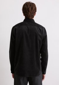 Mann mit kurzen braunen Haaren, der ein schwarzes Cordhemd mit langen Ärmeln trägt und mit dem Rücken zu einem schlichten hellen Hintergrund steht.