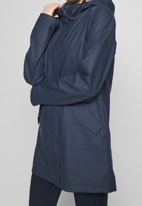 Mörkblå vattentät jacka med huva, utrustad med en främre dragkedja, tryckknappar och sidofickor. Slät textur och rak skärning.