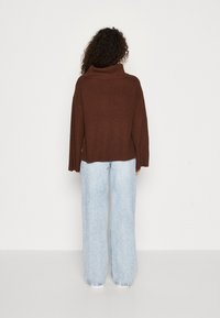 Suéter marrón de canalé con cuello tipo capucha y mangas anchas, combinado con jeans de pierna ancha en azul claro, visto desde la espalda sobre un fondo liso.