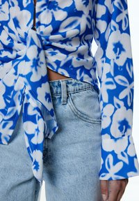 Blaue und weiße Bluse mit Blumenmuster und Krawatten-Design, kombiniert mit hellblauen Jeans. Strukturierter Stoff und ausgestellt geschnittene Ärmel.