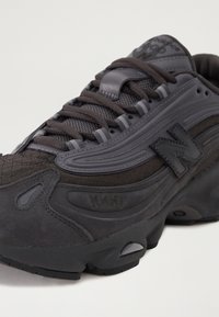 Baskets New Balance noires et gris foncé avec panneaux en mesh et en daim, semelle épaisse et fermeture à lacets, vues de côté.