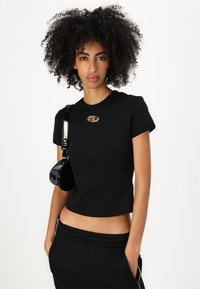 Svart avkortet t-skjorte med korte ermer, med et metallisk logo lapp. Kombinert med en liten svart patent crossbody-veske.