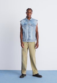 Denim ærmeløs jakke med to frontlommer i lys blå farve. Kombineret med olivengrønne bukser med sorte side-striber og sorte sko.