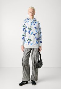 Hooded sweatshirt in white with colorful abstract figures and stars. Paired with loose gray cargo pants and black sneakers, holding a small bag.