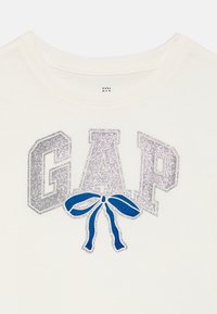 T-shirt couleur crème avec le texte "GAP" en paillettes argentées et un graphisme de nœud bleu en dessous des lettres. Tissu doux avec un col rond.