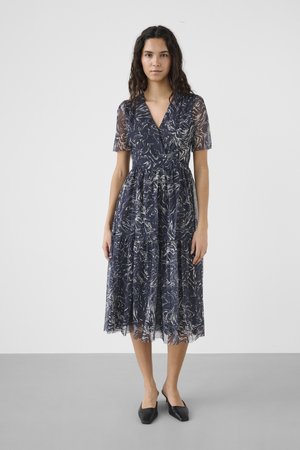 Moteris stovi, dėvinti tamsiai mėlynos ir baltos spalvų abstraktaus rašto midi suknelę su trumpomis rankovėmis ir juodus batelius su smailiais galiukais.