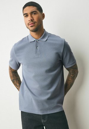 Camisa polo de manga corta azul claro con un tejido texturizado, con tapeta abotonada y cuello de canalé. Combinada con jeans oscuros.