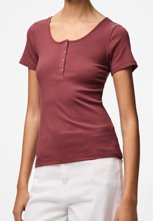 Femme portant un haut henley bordeaux ajusté à manches courtes côtelées, associé à un pantalon blanc taille haute, sur un fond uni.
