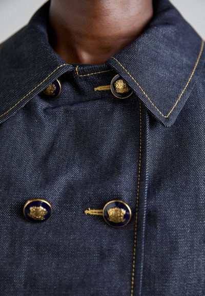 Temno modra denim jakna z širokim ovratnikom, zlatimi šivi in izrazitimi zlatimi gumbi s mornarsko modrim ozadjem.
