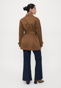 Brauner Wildledermantel mit Gürtel, Kragen, Rückenpasse und geknöpften Ärmelakzenten, kombiniert mit weit ausgestellten dunkelblauen Jeans.