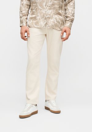 Mees, kes kannab beežikarva mustrilist nööbitavat särke, kreemikaarva pükse ja valgeid tenniseid pruunide talladega, seisab ühevärvilise tausta ees.