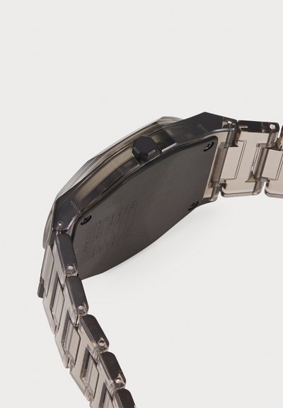 Dark gray watch with a rectangular case, transparent band links, and a prominent black crown. The back features printed technical details.