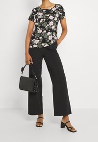 Top corto de flores negras con flores rosas y verdes, combinado con pantalones anchos negros. Sosteniendo un bolso negro texturizado y llevando sandalias negras con tiras.
