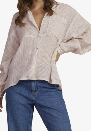Femme portant une chemise texturée rose clair à boutons avec des manches longues et un jean bleu, debout avec une main sur la hanche.