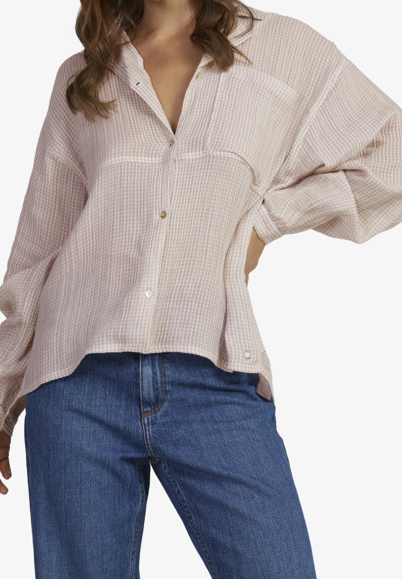 Femme portant une chemise texturée rose clair à boutons avec des manches longues et un jean bleu, debout avec une main sur la hanche.