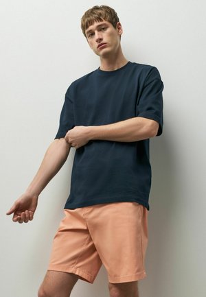 Junger Mann trägt ein dunkelblaues übergroßes T-Shirt und hellorangefarbene Shorts, zieht seinen linken Ärmel vor einem einfarbigen weißen Hintergrund zurecht.