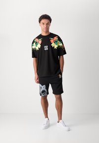 T-shirt noir surdimensionné avec un graphique floral sur les épaules, logo "HUGO" blanc ; associé à un short noir avec une impression sur le côté, porté avec des baskets blanches.