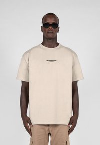 Beige oversized t-shirt med korte ærmer, der har et lille sort tekstlogo centreret på brystet. Båret med lysebrune cargo-bukser og mørke solbriller.