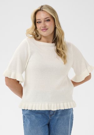 Žena s dugom plavom kosom koja nosi bijelu pletenu majicu s naboranim rukavima i rubom, u kombinaciji s plavim trapericama, na jednobojnoj pozadini.