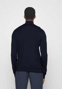 Pull en maille bleu marine avec un col montant, des manches longues, et des poignets et ourlet côtelés. Texture lisse, design minimaliste.