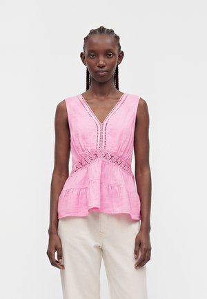 Vrouw met gevlochten haar, die een mouwloze roze V-hals top met kantdetails draagt en een witte broek met hoge taille tegen een effen achtergrond.