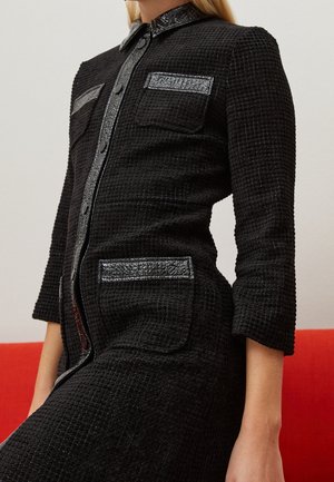 Czarna sukienka o fakturowanej tkaninie z dopasowanym krojem, zapięciem na guziki, metalicznymi detalami na kołnierzu i kieszeniach oraz rękawami o długości do łokcia.