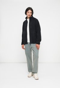 Giovane che sta in piedi indossando una giacca in pile nera con zip, una maglietta bianca, jeans azzurri chiari e sneakers bianche contro uno sfondo bianco semplice.