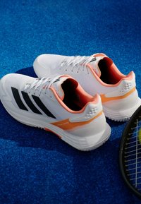 Λευκά αθλητικά παπούτσια με μαύρες ρίγες, πορτοκαλί λεπτομέρειες, πλεκτό υλικό, υφασμάτινες σόλες και συνολική αεροδυναμική σχεδίαση.