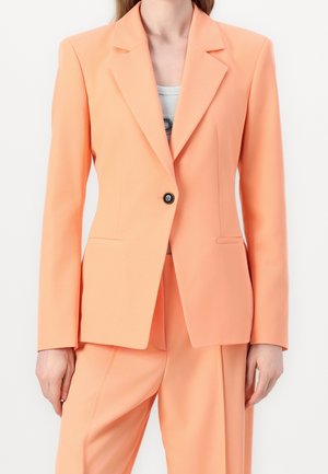 Kvinde iført en ferskenfarvet skræddersyet blazer med en sort knap, kombineret med matchende højtaljede bukser og en lys indertop.