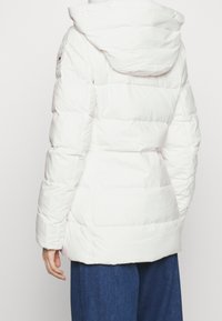 Vit puffjacka med huva, med kviltade sektioner och en logotyp på vänster axel. Mjuk, slät textur som ger värme.