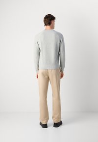Pull gris clair à côtes avec un col rond, porté avec un pantalon beige et des chaussures noires, vu de dos sur un fond uni.