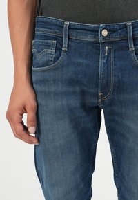 Sötétkék farmernadrág, slim fit szabással, klasszikus öt zsebes kialakítással, látható varrásokkal és fém gombos derékzárral.