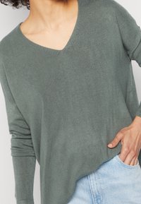 Grøn v-hals sweater med lange ærmer, strikket tekstur, afslappet pasform og buet kant, parret med lyseblå jeans.