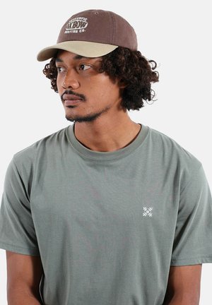 Homme aux cheveux bouclés portant un t-shirt vert avec un petit logo blanc et une casquette marron-beige, regardant vers la gauche sur un fond uni clair.