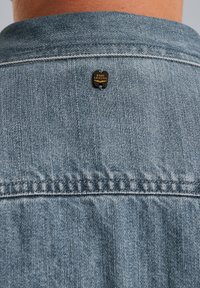 Jasnoniebieska dżinsowa kurtka z klasycznym kołnierzem, z czarną i złotą metką marki wszytą w okolicy tylnej części dekoltu.