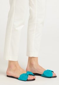Sandale din suede turcoaz cu vârf deschis, decorate cu o fundă mare, având talpa plată de culoare neagră, purtate cu pantaloni albi la dungă.