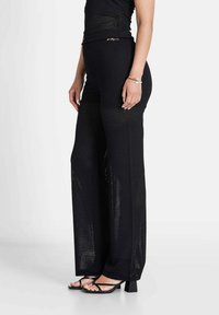 Zwarte wijde broek gemaakt van textuur, open breiwerk met zijsplits, gecombineerd met zwarte sandalen met bandjes en eenvoudige accessoires.