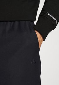 Main dans la poche d'un pantalon de survêtement noir Calvin Klein avec le logo « cK » brodé, portant un sweat-shirt noir avec le logo Calvin Klein sur la manche.
