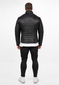 Zwart leren jack met een gequiltte textuur, hoge kraag en een hexagonaal patroon op de rug. Gedragen over een wit shirt met zwarte jeans en sneakers.