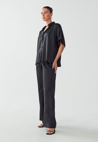 Camicia di satin nera con maniche corte e bottoni, abbinata a pantaloni larghi in satin coordinati. Design semplice ed elegante con struttura liscia.