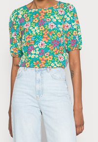 Blouse à imprimé floral avec un fond vert vif, des manches courtes bouffantes et une coupe décontractée ; associée à un jean taille haute bleu clair.