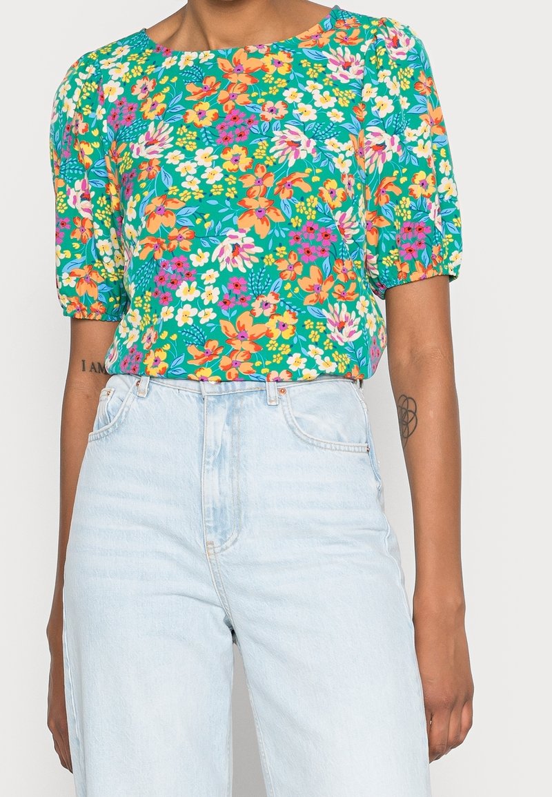 Blouse à imprimé floral avec un fond vert vif, des manches courtes bouffantes et une coupe décontractée ; associée à un jean taille haute bleu clair.