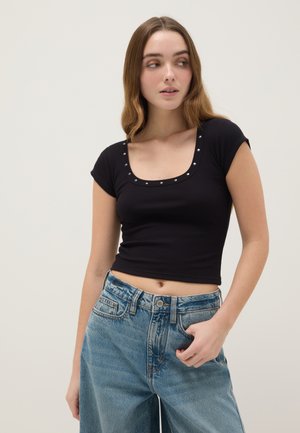 Jeune femme aux cheveux longs et ondulés portant un crop top noir à encolure dégagée orné de clous et un jean bleu taille haute, regardant sur le côté.